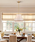 3 Light Gold Pendant Light Featuring White Linen Cone Shade And Sturdy Metal Cage hanging above a dining table in a farmhouse setting.