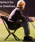Person sitting on an Ultra Compact Portable Camp Chair That Folds To Bottle Size Ready In Seconds A Real Game Changer For Camping And Outdoors Supports Up To 350 Lbs Lightweight And Built For Hiking Adventures Black, on a field sideline at night.