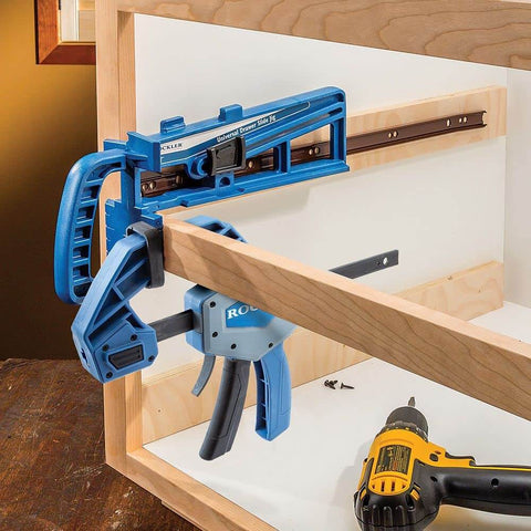 Universal Drawer Slide Jig For Cabinets Easy Install Drawer Slides Quickly And Accurately With A Locking Wedge Adjustable Indexing Foot And Large Clamping Area For Pro Grade Cabinet Hardware Installations with blue tool clamping on wooden cabinet frame.