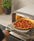 Ooni 12-inch aluminum pizza peel with handle inserting pepperoni pizza into a compact countertop oven