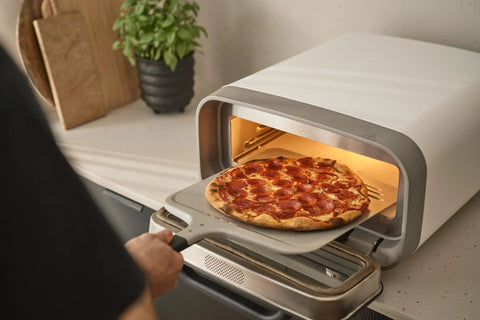 Ooni 12-inch aluminum pizza peel with handle inserting pepperoni pizza into a compact countertop oven