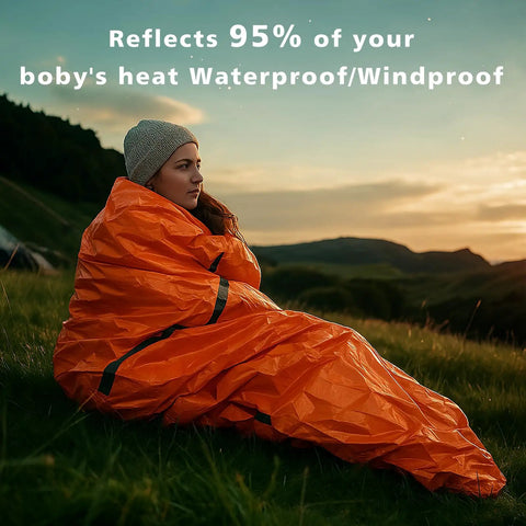 Person in bright orange Emergency Mylar Sleeping Bag Set For Camping Waterproof Thermal Bivy Sacks outdoors at sunset.