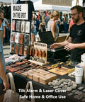 Market stall displaying leather wallets, wooden coasters, and engraved keychains on a table; vendor uses a laptop.