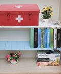 Red metal first aid kit box with white cross emblem and two latches on a white shelf.