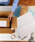 Person using 100 Sheets Newsprint Packing Paper for Moving and Shipping on a table with packing materials and cardboard box