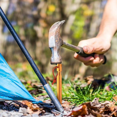 Hammer driving 12 Pack Tent Stakes Made From 7075 Aluminum into ground for stability, showing ultra-lightweight orange peg use in camping.