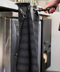 Top Shelf Espresso Knock Box Built Different For Quick Grounds Ejection Splash Proof Tilt Design Heavy Duty Stainless Steel Build Removable Base And Barista Towel hanging on a kitchen counter.