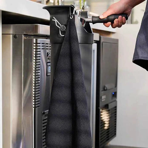 Top Shelf Espresso Knock Box Built Different For Quick Grounds Ejection Splash Proof Tilt Design Heavy Duty Stainless Steel Build Removable Base And Barista Towel hanging on a kitchen counter.