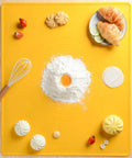Yellow baking mat with flour, egg yolk, whisk, tomatoes, pastries, and dumplings on wooden table