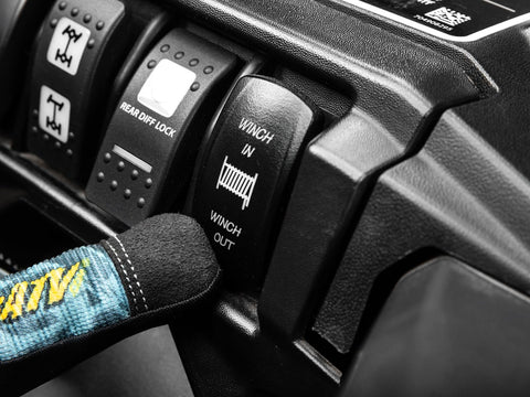 Close-up of a black UTV winch control panel labeled WINCH IN and WINCH OUT, with a blue strap in the foreground.