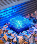 Blue illuminated solar glass brick path light nestled in gravel, part of Ice Cube Solar Glass Brick Path Lights 4 Pack For Garden And Yard.