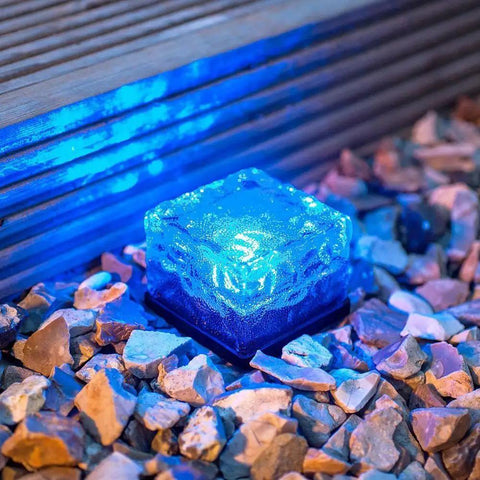 Blue illuminated solar glass brick path light nestled in gravel, part of Ice Cube Solar Glass Brick Path Lights 4 Pack For Garden And Yard.
