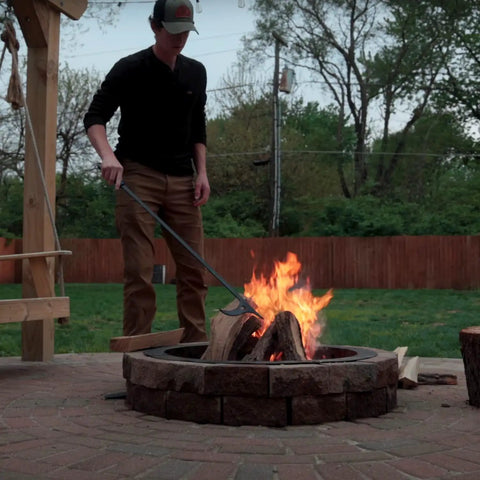 Person using 40 Inch Fire Poker And Log Grabber Set For Fire Pits Must Have Backyard Outdoor Indoor Bonfire Gear Rustproof Two Piece Campfire Tool With Ergonomic Rubber Handles For Safe Easy Firewood Movement on a stone fire pit in a backyard.