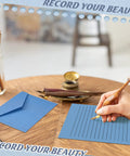 Hand writing on lined paper with pen beside blue envelope, part of the All In One Blue Letter Writing Paper And Envelope Set With 50 Lined Papers 50 Envelopes And 50 Stickers Includes A Safe Box For Easy Storage Sized 5.5 X 8.25 Inches Great For Office Notes Personal Letters And School Projects.