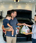 Family unloading groceries from a car trunk, featuring a child in the father's arms.