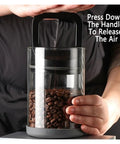 Premium Vacuum Sealed Clear Glass Coffee Bean Canister With Secure Airtight Lid To Keep Kitchen Food Storage And Tea Moisture Proof While Locking Down Freshness For Your Daily Grind In Sleek Black with beans inside and hand pressing the air release handle