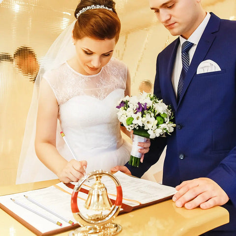 Bride and groom signing a wedding registry at a decorated table, with the bride holding a bouquet and groom in a blue suit.