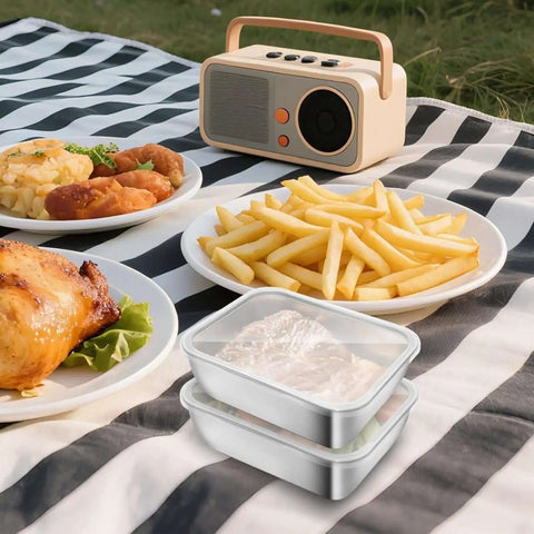 Two stainless steel storage containers with clear lids on a picnic blanket with food and a retro radio