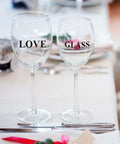 Two clear wine glasses with black LOVE and GLASS lettering on the bowls, placed on a dining table with cutlery.