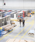 Interior of a warehouse with workers walking, surrounded by shelving units and heavy duty steel casters on carts for industrial use.