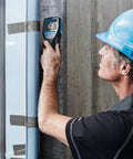 A man uses The Real Deal Digital Wall Scanner And Floor Detection Tool on a concrete wall to locate hidden wires and pipes.