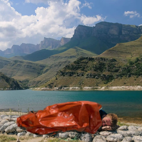 Person in an Emergency Survival Sleeping Bags 2 Pack For Cold Weather Camping And Outdoor Rescue Mylar Thermal Bivy Sack by a lake