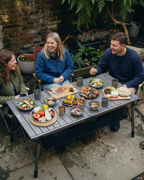People enjoying a meal on a 50 Inch Camping Table With Adjustable Height Legs For Uneven Ground, Easy Setup Portable Folding Aluminum Roll-Up Table With Mesh Pocket For Camping, Picnic, BBQ, Beach And Outdoor Cooking.