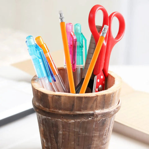 Wooden cup holding assorted pens, scissors, and pencils, including a yellow carpenter pencil with white eraser tips for construction