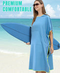 Woman at the beach in blue hooded towel changing poncho for surfing and outdoor adventures, holding a surfboard.
