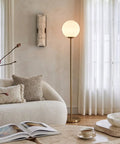 Luna LED Floor Lamp Dope Modern Tall Light For Living Rooms And Home Offices With Frosted Glass Globe Mid Century Style Standing Lamp In Brass Gold Finish Perfect For Reading In The Bedroom And Cozy Vibes next to a sofa in a cozy living room.