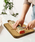 Acacia wood cutting board with handle on a white countertop; person slices a sprinkles-covered rolled cake.