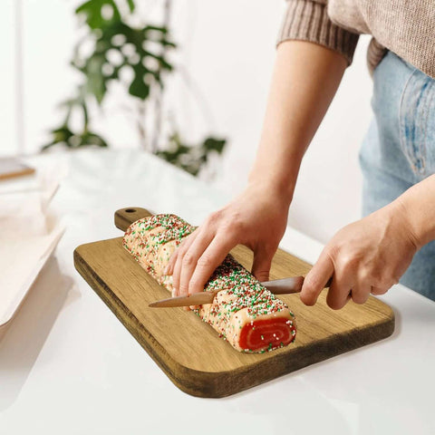 Acacia wood cutting board with handle on a white countertop; person slices a sprinkles-covered rolled cake.