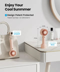 Mini Portable Handheld Fan With Base 3 Speed USB Rechargeable Dual Use Desk And Personal Fan Pink on table next to bag