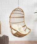 Mariah Wicker Hanging Chair With Cushion For Indoor Outdoor Chill Lounger, shown against a white brick wall in light brown and beige.