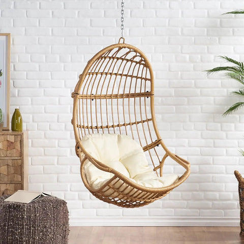 Mariah Wicker Hanging Chair With Cushion For Indoor Outdoor Chill Lounger, shown against a white brick wall in light brown and beige.