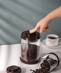 Round High Borosilicate Glass Coffee Bean Container With Real Deal Airtight Seal showcasing coffee beans and a hand pressing the lid.