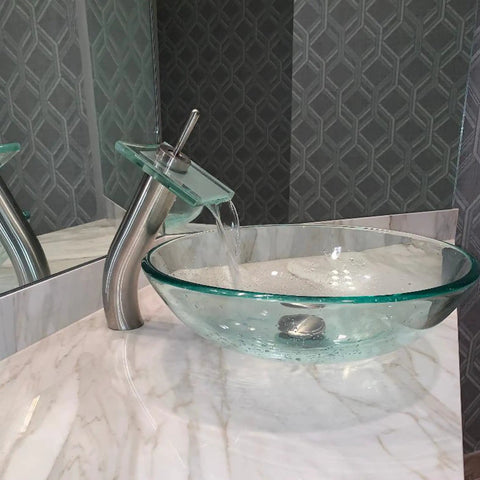 Squared Single Lever Waterfall Vessel Faucet In Brushed Nickel With Clear Glass, next to round glass vessel sink on marble countertop