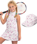 Girl wearing pink girls athletic dress with UPF 50 plus sun shield, matching hat, two side pockets, bow accent, sleeveless golf and tennis gear, ages 4 to 5, easy move comfort, holding tennis racket.