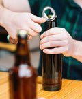 Ring-style metal bottle opener in use to remove a cap from a brown beer bottle; second bottle visible in the foreground.