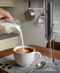 Rechargeable Handheld Milk Frother With Stand For Coffee Lattes Cappuccinos Matcha And More Quiet Fast Foam Wand In 15 To 30 Seconds USB Rechargeable Kitchen Gift Black next to cup of coffee being prepared on kitchen table.