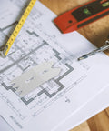 Close-up of a drill bit, tape measure, and level on architectural plans, unrelated to Set Of Four Heavy Duty Bench Dog Clamps For Woodworking.