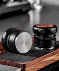 Two coffee tampers on a wooden mat, featuring a round steel base and ergonomic design, part of the Slick Espresso Tamper Set For 58Mm Portafilters With Auto Scale Dial Wdt Tool And Smart Storage Mat A Fire Four Piece Kit That Straight Up Upgrades Your Daily Coffee Grind Game.