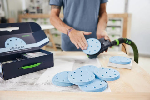 Person using Granat D125 GR sanding discs for VOC varnishes, showing clog-resistant discs and packaging on a workspace.
