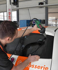 Green hot air welding gun used on car windshield by technician in auto workshop; power cord visible.
