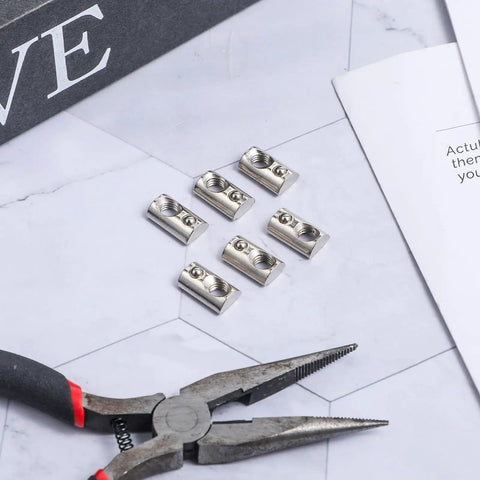 Close-up of 6 roll-in T-nuts with ball spring for 10 series aluminum extrusion profiles, 1/4-20 size, zinc plated steel on table with pliers