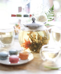 All In One Glass Teapot And Kettle With Heatproof Borosilicate Glass Removable Stainless Steel Infuser Microwave And Stovetop Friendly Tea Maker For Everyday Tea Lovers 40 Oz 1200 Ml with blooming tea, surrounded by elegant pastries and flowers on a wooden table.