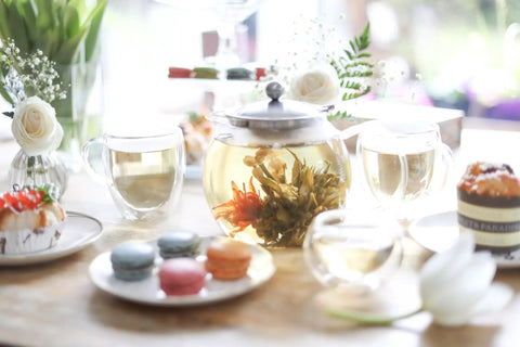 All In One Glass Teapot And Kettle With Heatproof Borosilicate Glass Removable Stainless Steel Infuser Microwave And Stovetop Friendly Tea Maker For Everyday Tea Lovers 40 Oz 1200 Ml with blooming tea, surrounded by elegant pastries and flowers on a wooden table.