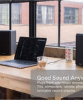 PB42X Powered Bookshelf Speakers With 30 Watt RMS beside a laptop on a wooden desk near large windows