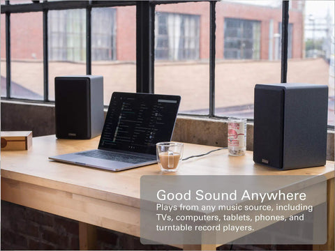 PB42X Powered Bookshelf Speakers With 30 Watt RMS beside a laptop on a wooden desk near large windows