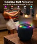 Room 3 Portable Bluetooth Speaker With IPX6 Waterproof Build TWS Pairing 12W Bass Boomin Stereo Sound With RGB Lights Bluetooth Five Four Ideal For Any Surface At Home on a table with RGB lights glowing, ambient setting in background.